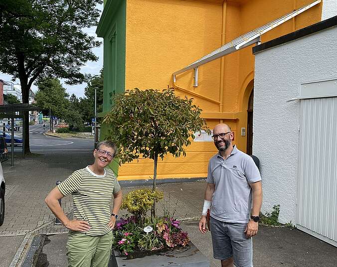 Das Foto zeigt links die Museumsleiterin Friederike Lutz mit dem Chef des Blumenhauses Mayer, Florian Mayer. Sie stehen neben einem Pflanzgefäß, das Herr Mayer dem Schulmuseum ausleiht.