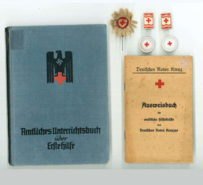 Ein blaues Buch mit dem Titel "Amtliches Unterrichtsbuch über Erste Hilfe", ein hellbraunes "AUsweisbuch für weibliche Hilfskräfte des Deutschen Roten Kreuzes" und fünf Abzeichen des WInterhilfswerk, die alle das Logo des Roten Kreuzes tragen, liegen nebeneinander.