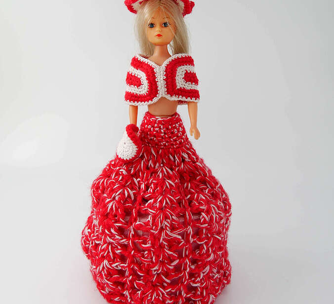 Eine Barbiepuppe in einem roten gehäkelten Kleid. Unter Ihrem Rock ist eine Rolle pinkfarbenes Klopapier.