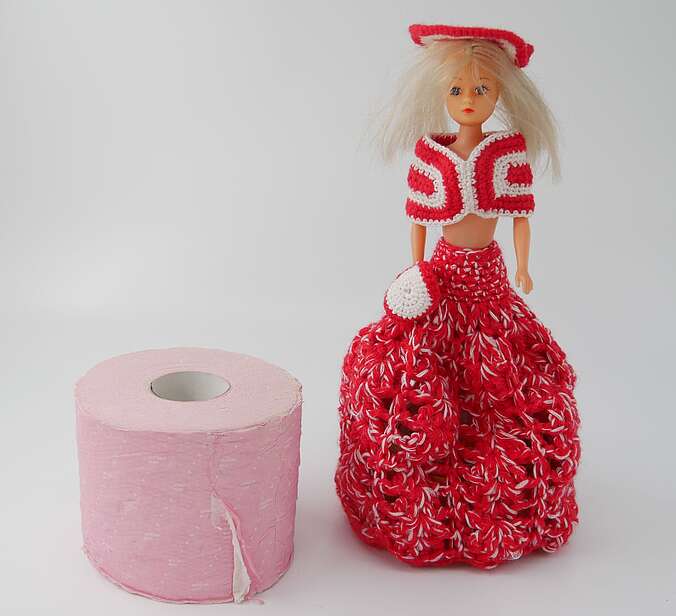Eine Rolle pinkfarbenes Klopapier steht neben einer Barbiepuppe, die ein rotes gehäkeltes Klied trägt.