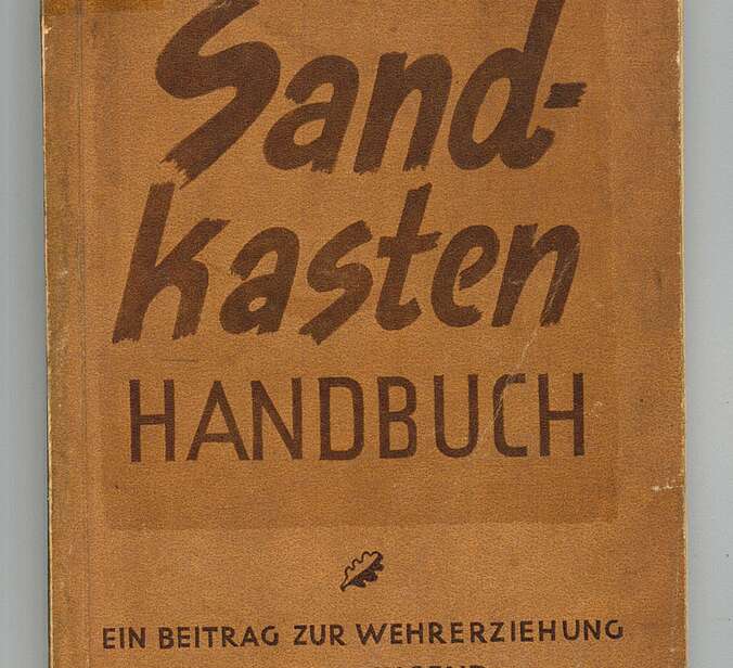 Die Titelseite eines braunen Buches. In dunkelbraunen Buchstaben steht darauf "Sandkastenhandbuch. Ein Beitrag zur Wehrerziehung der Schuljugend."